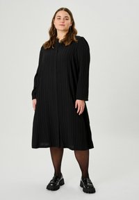 Schwarzes, plissiertes Kleid mit langen Ärmeln und Knopfverschluss. Gerundeter Halsausschnitt und gewellte Bündchen. Getragen mit glänzenden schwarzen Plateauschuhen.