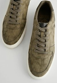Maslinasto zelene suede tenisice s teksturiranom površinom, zaobljenim vrhom i bijelom gumenom potplatom. Imaju ravne vezice i suptilne šavove.