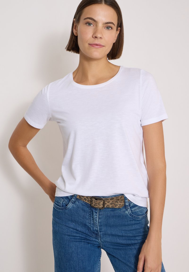 Femme aux cheveux courts bruns portant un t-shirt blanc uni, un jean bleu et une ceinture marron à motif léopard, debout devant un fond clair.