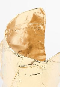 Polsino metallico dorato con superficie texture, caratterizzato da bordi frastagliati e sottili imperfezioni, che ne sottolineano la qualità artigianale e la finitura riflettente.