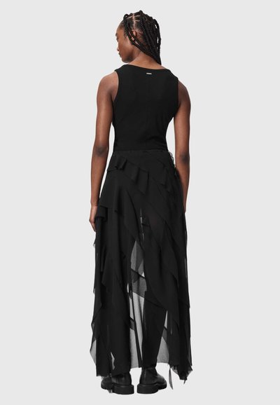 Femme aux cheveux tressés, portant un haut noir sans manches et une jupe en maille noire superposée et asymétrique avec des bottes noires, vue de dos.