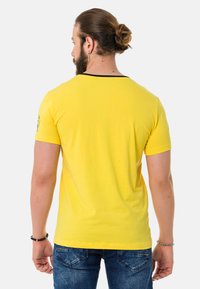 Gelbes T-Shirt mit kurzen Ärmeln und schwarzem Ausschnitt, glatter Textur und einem Emblem am linken Ärmel, kombiniert mit blauen Jeans.