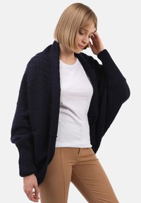 Marinblå stickad cardigan med strukturerade mönster, �öppen fram och ballongärmar. Kombinerad med en vit topp och beige byxor för kontrast.