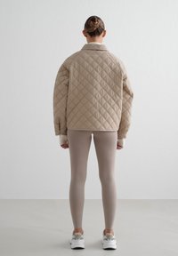 Beige quiltet jakke med en avslappet passform, kombinert med lyse beige leggings. Jakken har krage og en merkbar diamantmønster. Hvite sneakers fullfører antrekket.