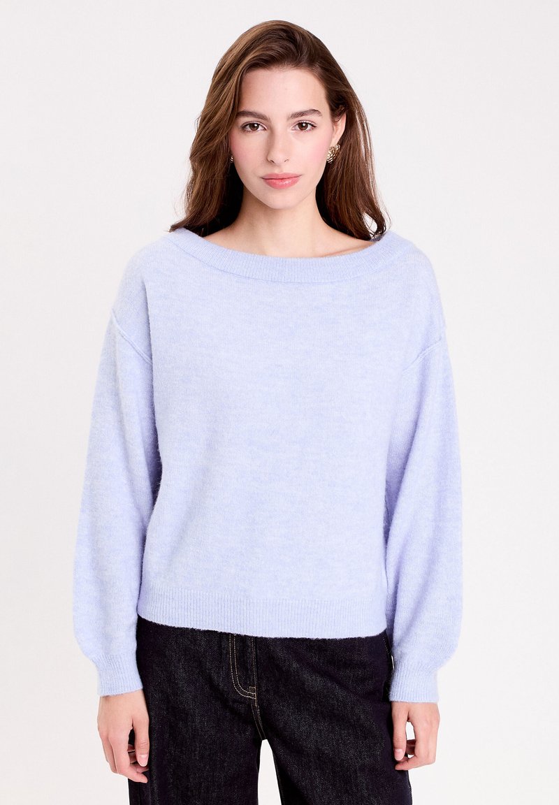 Sweater tricoté bleu clair avec un large col bateau, des épaules tombantes, des poignets et un ourlet côtelés. Tissu doux et texturé ; associé à un jean foncé.