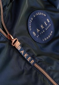 Giacca navy con una texture liscia, dotata di un logo in tessuto rotondo e una zip in rosé dorato con dettagli coordinati. Etichetta "BAKER" sul nastro della zip.