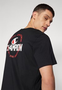 Sort bomulds T-shirt med et stort rundt rødt logo og "CHAMPION" tekst på ryggen, korte ærmer og klassisk crew neck design.