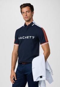 Marineblauwe polo met witte en oranje accenten, met het "HACKETT" logo in het wit. Gepaard met marineblauwe broeken en een witte sweatshirt in de hand.