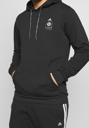 Felpa nera con cappuccio e tasca frontale, caratterizzata dal logo Adidas e da stringhe bianche. Tessuto morbido, vestibilità casual e polsini a coste.