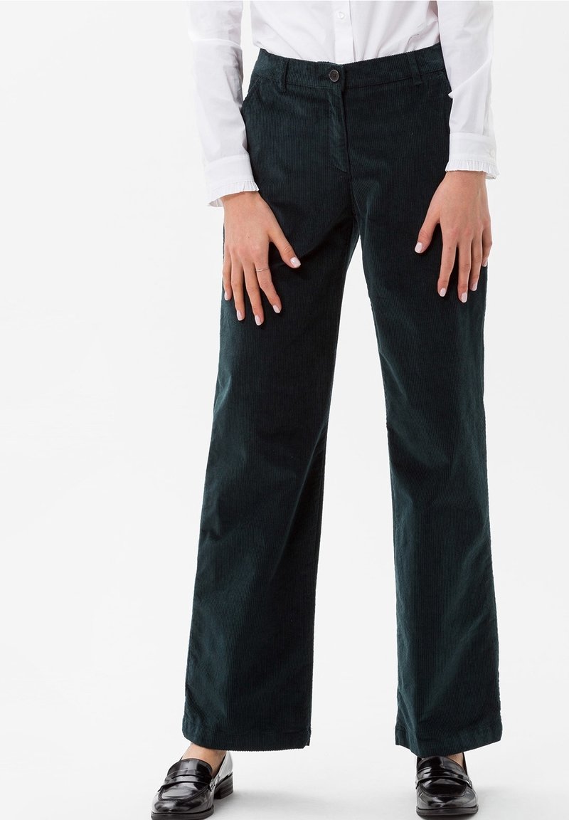 Femme portant un pantalon large en velours côtelé noir, une chemise blanche et des mocassins noirs, debout avec les mains détendues le long du corps.