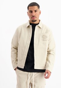 Beige corduroy jack met drukknoopsluiting, borstzak met geborduurd logo, gedragen over een zwart shirt, textuurstof.