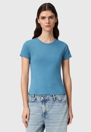 Ung kvinde med langt brunt hår iført en tætsiddende himmelblå kortærmet t-shirt og lyseblå højtaljede jeans, stående foran en ensfarvet baggrund.