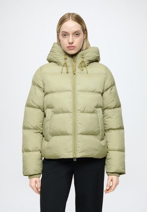 Donna che indossa una giacca imbottita verde chiaro con grande cappuccio e pantaloni neri, in piedi davanti a uno sfondo chiaro uniforme.