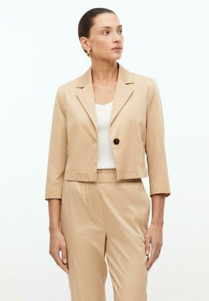 Vrouw die een beige cropped blazer met één knoop draagt, bijpassende hoge taille broek en een wit topje, kijkt opzij tegen een witte achtergrond.