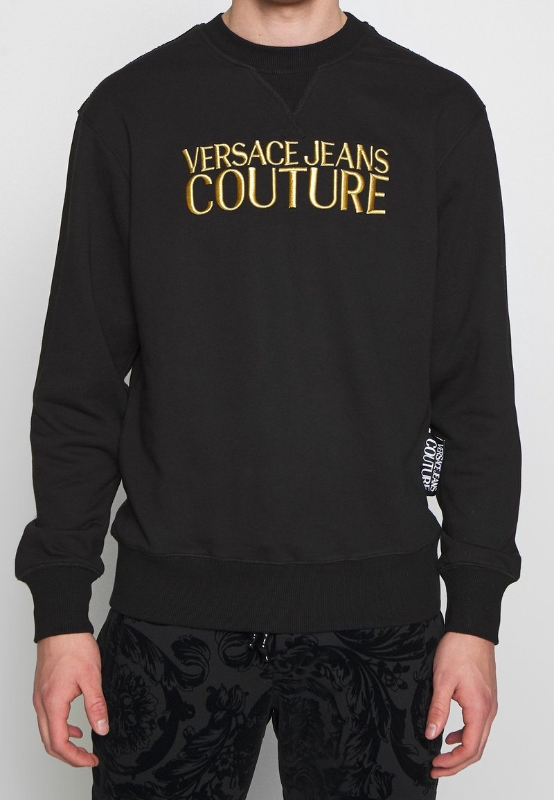 Personne portant un sweat-shirt noir avec le texte doré « Versace Jeans Couture » sur la poitrine et un pantalon noir à motif floral.