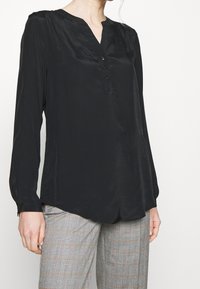 Kvinna klädd i en svart långärmad blus med knappar och grå rutiga byxor med svaga röda och orangea linjer.