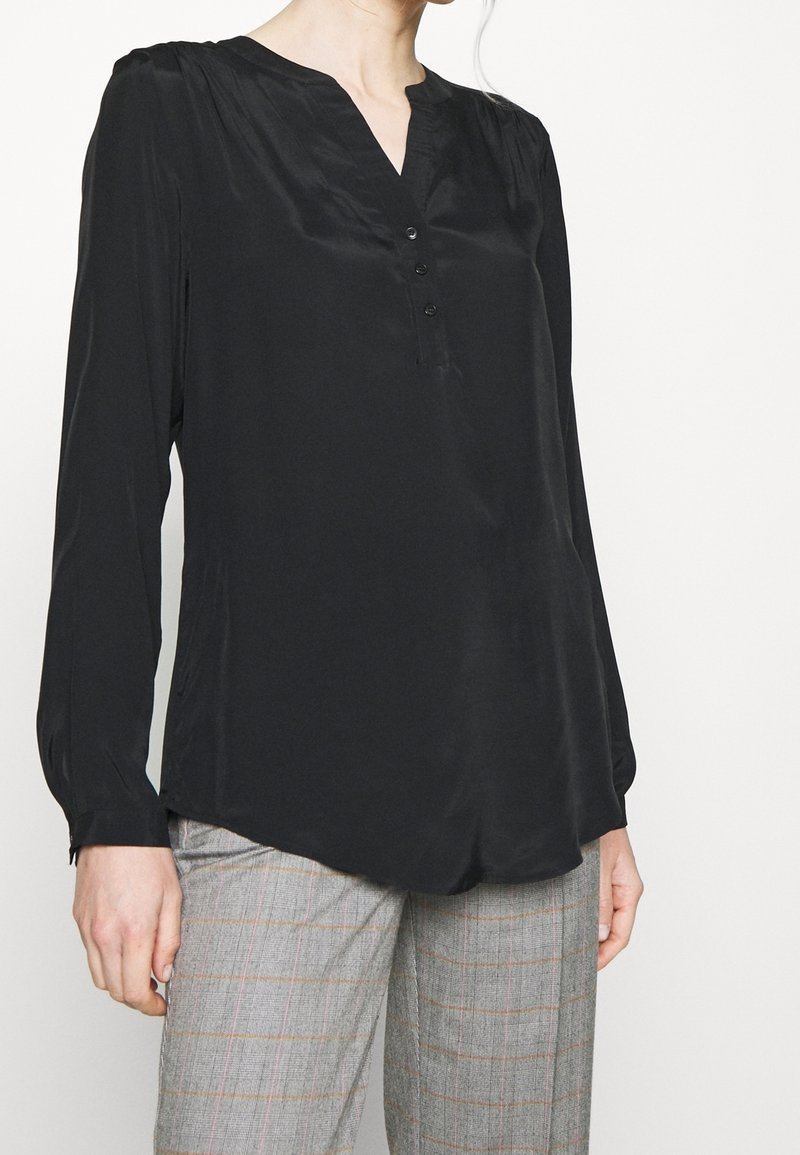 Kvinna klädd i en svart långärmad blus med knappar och grå rutiga byxor med svaga röda och orangea linjer.