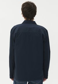 Moški s kratkimi rjavimi lasmi, oblečen v temno mornarsko srajco z dolgimi rokavi in modre kavbojke, viden s hrbta proti enotnemu svetlemu ozadju.