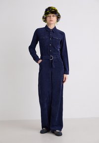 Marineblauwe corduroy jumpsuit met lange mouwen, knoopsluiting aan de voorkant, twee borstzakken en een tailleband. Gecombineerd met een kleurrijke, pluizige hoed.