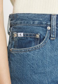 Blue denim jeans feature a visible front pocket with a white CK label, silver rivets, and contrasting gold stitching along the seams.