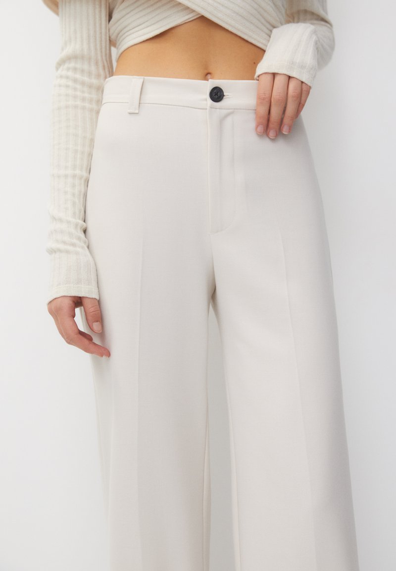 Pantaloni beige chiaro su misura con gamba dritta, piega frontale e chiusura con un unico bottone scuro. Texture liscia, design minimalista.