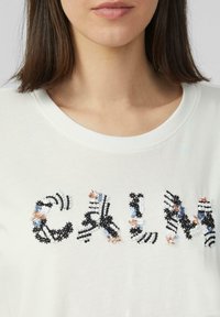 Weißes Baumwoll-T-Shirt, das das Wort "CALM" in schwarster, blauer und rosa, floraler Stickerei mit Pailletten und Streifen auf einer strukturierten Oberfläche zeigt.