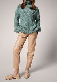Teksturowany zielony sweter ze żebrowanym wzorem i wysokim kołnierzem, zestawiony z beżowymi spodniami z sztucznej skóry oraz brązowymi mokasynami na grubej podeszwie.