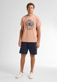Camiseta de algodón rosa claro con un diseño gráfico azul marino que presenta un sol y texto. Combinada con pantalones cortos azul marino oscuro y zapatillas blancas.
