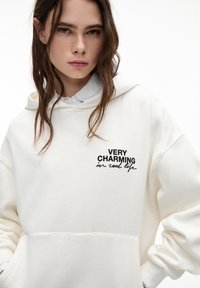 Hoodie surdimensionné blanc avec le texte noir "TRÈS CHARME EN VRAI." Comprend une poche kangourou et un capuchon avec cordon de serrage. Texture douce.