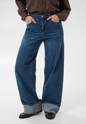 Personne portant un jean bleu à jambes larges avec de grands revers, une chemise marron boutonnée et des chaussures marron, debout les mains dans les poches.