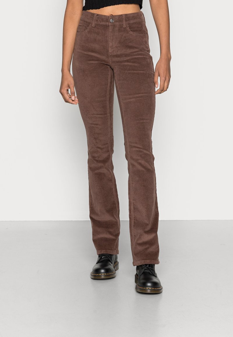 Pantalon en velours côtelé marron avec une coupe ajustée, présentant une fermeture à bouton, cinq poches et une surface nervurée texturée. Porté avec des chaussures noires.