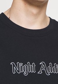 Černé bavlněné tričko s kulatým výstřihem, na přední straně s bílým stylizovaným textem "Night Add". Hladký povrch, záběr zblízka.