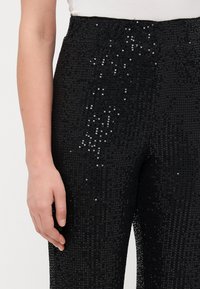 Czarne spodnie z cekinami z wysokim stanem, o fakturowej powierzchni pokrytej małymi okrągłymi cekinami, odbijającymi światło. Zbliżenie na rękę spoczywającą na tkaninie.