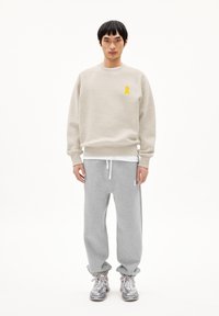 Man die naar voren kijkt, gekleed in een beige sweatshirt met een geel logo, grijze sweatpants met witte trekkoorden en zilveren sportSneakers.
