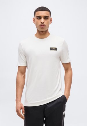 Hombre con camiseta blanca EA7 y pantalones negros, de pie contra un fondo blanco liso, posando con una mano en el bolsillo.