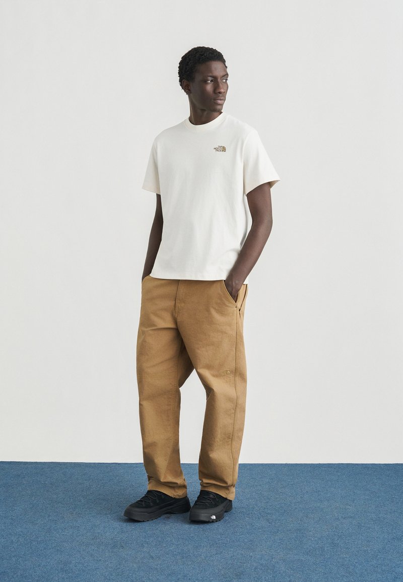 T-shirt en coton blanc avec petit logo, associé à un pantalon cargo marron clair. Baskets noires avec détails texturés. Fond plat et minimaliste.