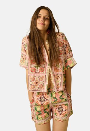 Jonge vrouw met lang bruin haar, gekleed in een kleurrijk patroon kort mouw shirt en bijpassende shorts, handen in zakken, neutrale achtergrond.