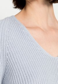 Ljust grå stickad tröja med djup V-ringning och texturerat mönster, som framhäver den ribbade ytan och det mjuka garnmaterialet.