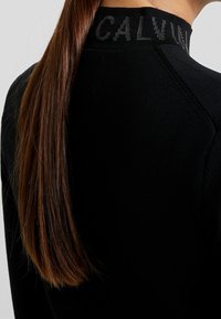 Top noir à manches longues avec col haut portant l'inscription "CALVIN", texture côtelée, design ajusté et coutures d'épaule raglan subtiles.