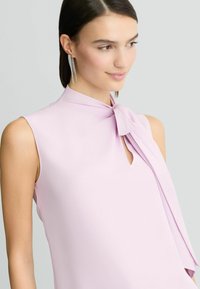 Lavendelkleurig mouwloos topje met een strikdetail bij de hals en een subtiele sleutelgatopening. Gladde stoftextuur met geplooide accenten.