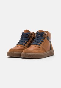 Baskets montantes brunes en cuir et daim, avec des lacets bleus, une fermeture éclair sur le côté, un bout perforé et une semelle en caoutchouc texturé.