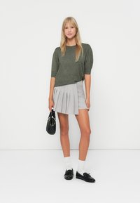 Un pull court à manches courtes de couleur vert olive associé à une mini-jupe grise plissée. Le mannequin tient un sac noir ; des chaussettes blanches et des mocassins noirs viennent parfaire le look.