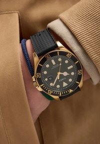 Montre dorée avec un bracelet en caoutchouc texturé noir, un cadran noir, des accents dorés et des marqueurs luminescents ; superposée à un bracelet en tissu bleu et vert.