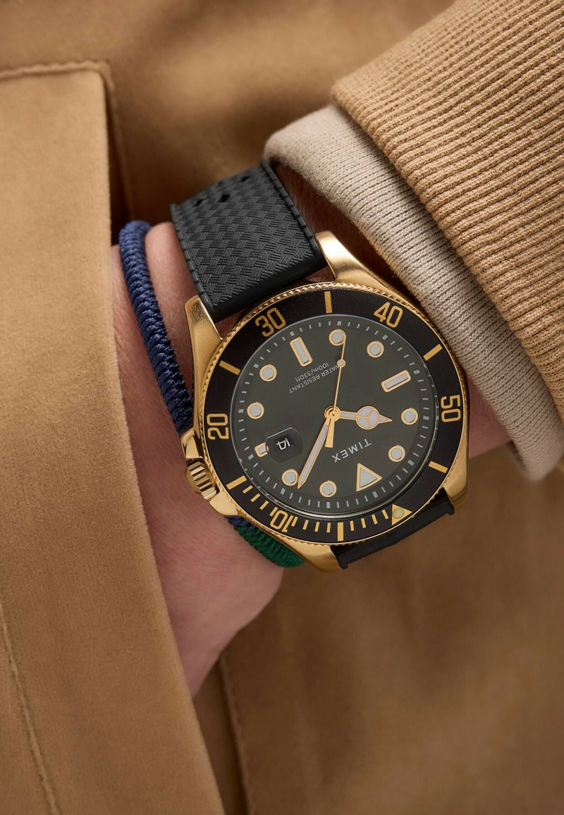 Orologio dorato con cinturino in gomma testurizzata nera, quadrante nero, dettagli dorati e indicatori luminosi; sovrapposto a un braccialetto in tessuto blu e verde.