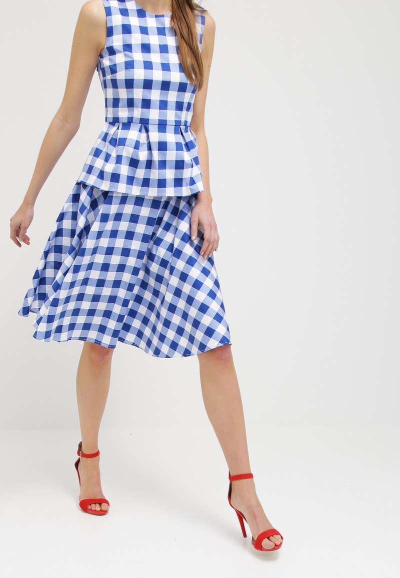 Blau-weißes Karomuster-Peplumkleid mit einem ausgestellten Rock. Kombiniert mit roten, spritzigen High-Heels mit Riemchen. Glattes Material mit einem schichtweisen Design.