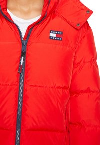 Red puffer jacket with a zip front, a hood, and a "TOMMY JEANS" logo patch. Textured fabric with a shiny finish; features ribbed trim.