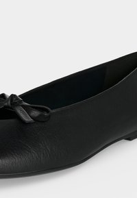 Ballerine plate en cuir noir avec bout arrondi et petit nœud décoratif à l'avant, présentée sur fond blanc.