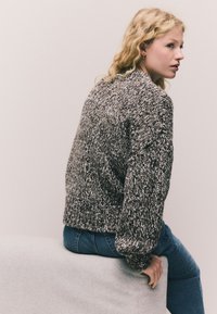 Pulkainsmēta džemperis melnā, baltā un pelēkā toņos. Raksturīgas plašas balona piedurknes un ribotas aproces. Valkāts ar zilām džinsa biksēm.