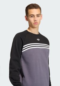 Crna i siva dukserica s bijelim horizontalnim prugama. Ima okrugli izrez i Adidas logo na prsima. Soft, teksturirani materijal.