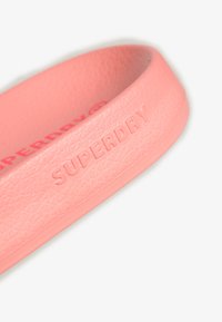 Gros plan sur une sangle de sandale en caoutchouc rose texturé, embossée avec le nom de la marque « Superdry » en lettres majuscules.